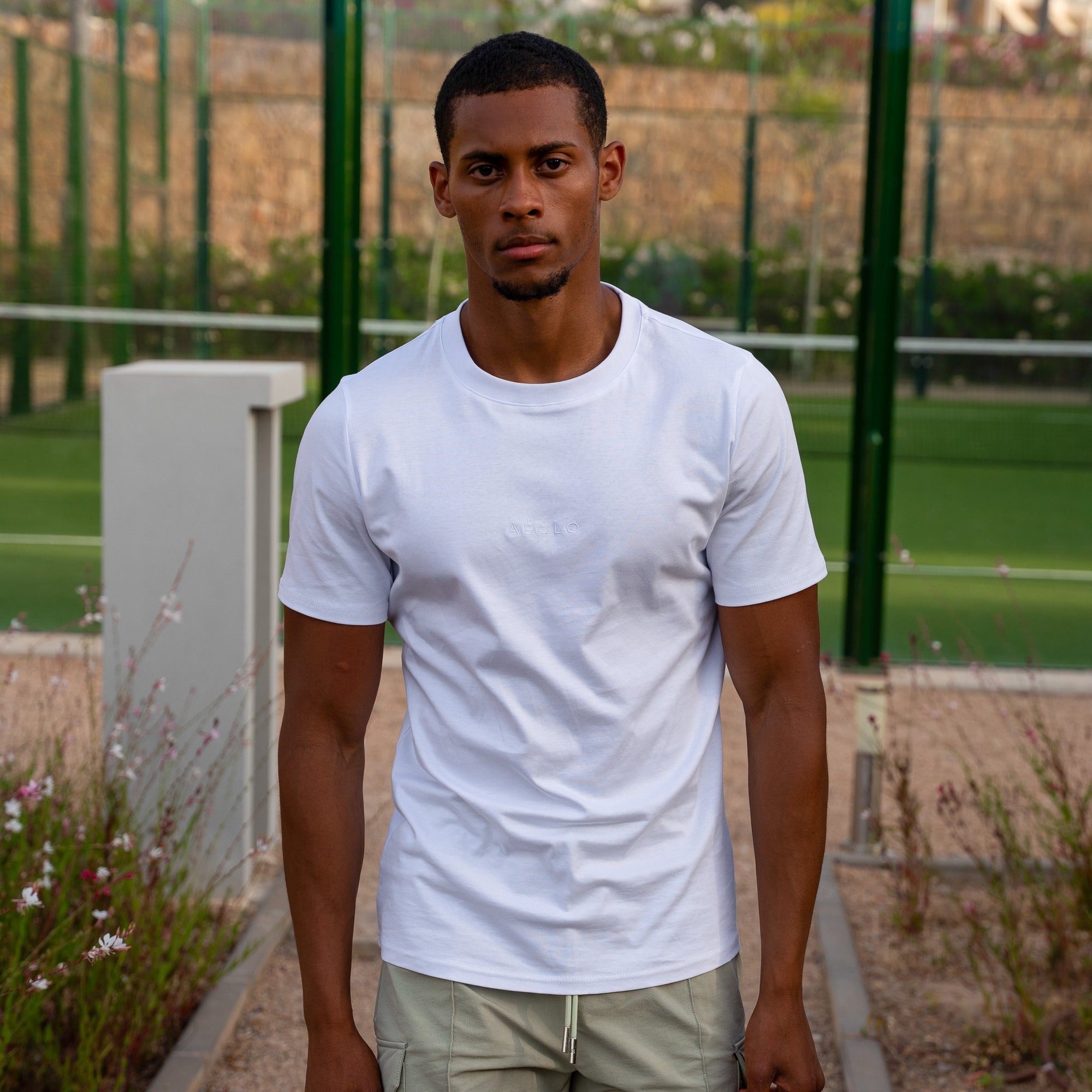 Man wearing a white t-shirt standing outdoors with greenery in the background